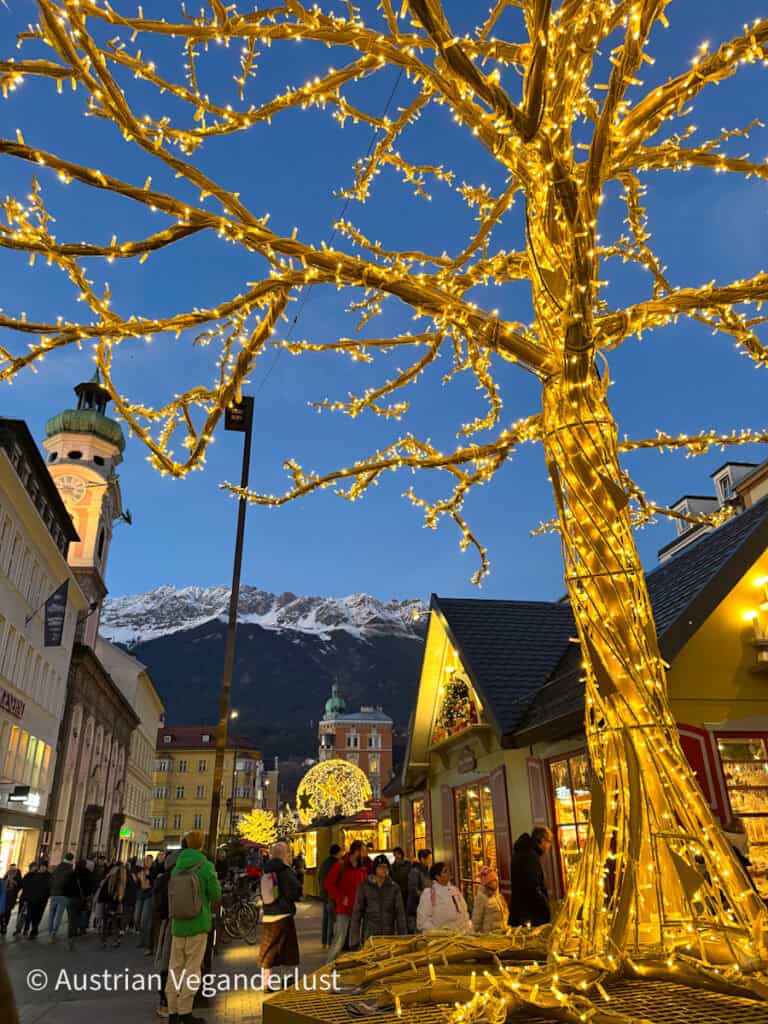 A golden light-wrapped artificial tree dominates the foreground of a festive Innsbruck street, with more decorations and snow-capped mountains completing the magical winter evening scene.