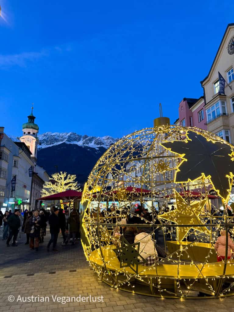 A large illuminated ornament structure with golden fairy lights and a bench inside, set along a festive pedestrian street in Innsbruck at twilight with the Nordkette mountains in the background.