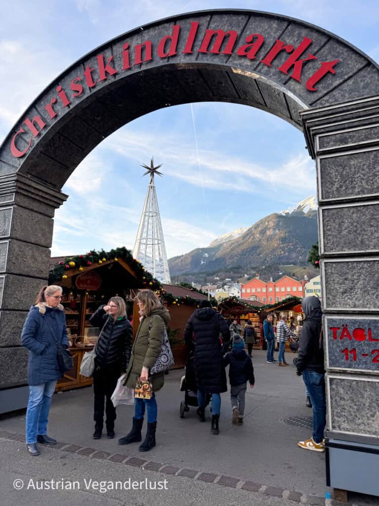 The stone archway entrance to the Christkindlmarkt with bold red lettering, opening onto rows of wooden stalls and a tall modern Christmas tree, with snow-capped mountains in the distance.