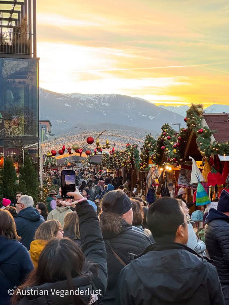 A crowded Christmas market lane at sunset, packed with people browsing decorated wooden stalls under garlands, with festive lights and mountain silhouettes in the background.
