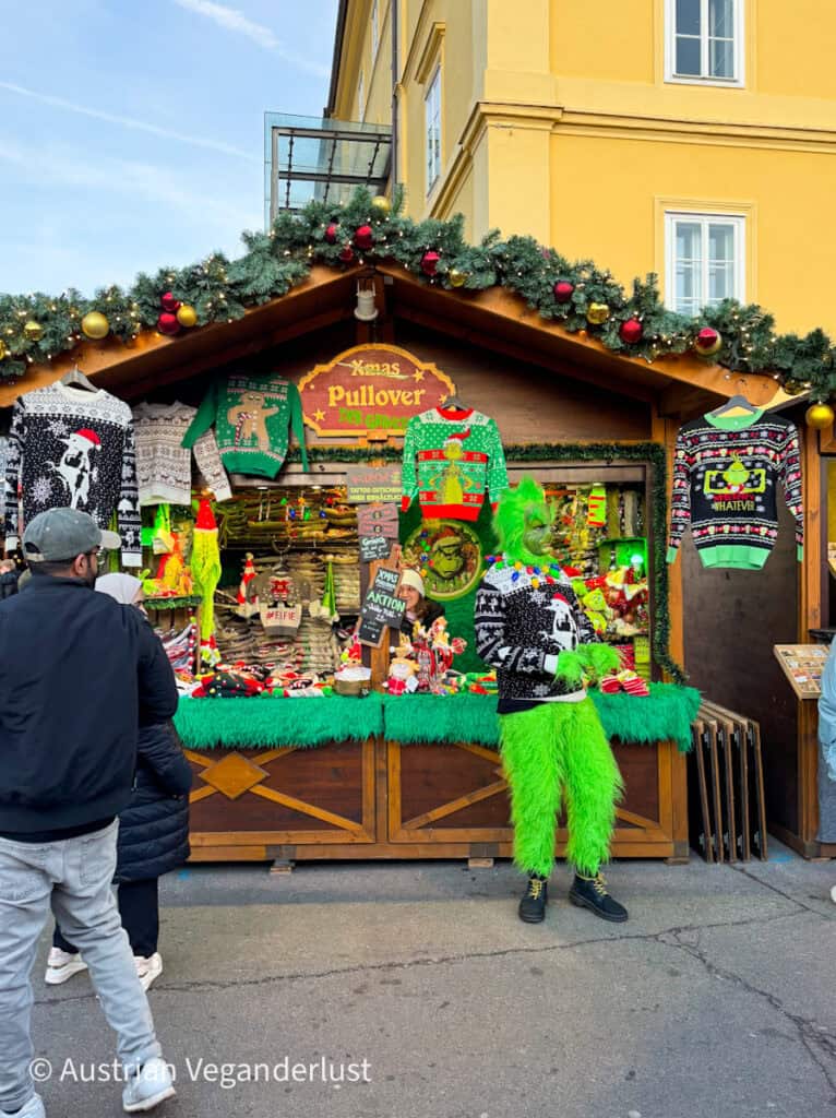 Christmas market stall selling novelty holiday sweaters, including one with a dancing gingerbread and another with “#WHATEVER” text. A person in a Grinch costume poses in front, matching the bright green decor.