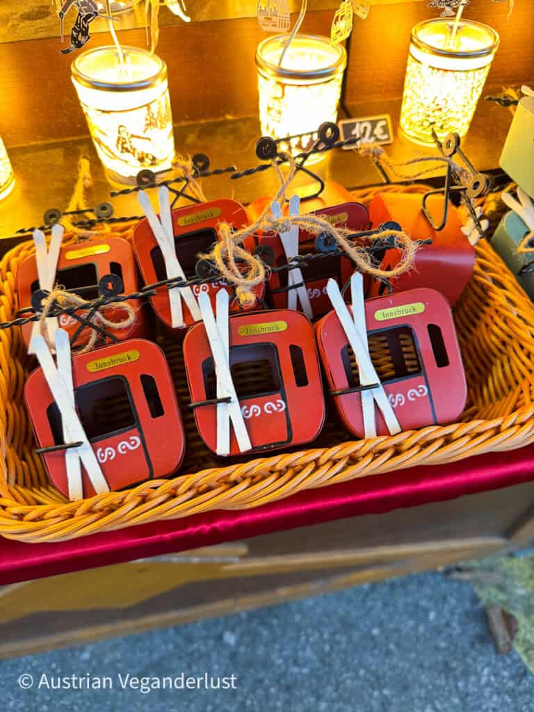 A basket of miniature red cable car ornaments labeled "Innsbruck" with white skis tied on top, displayed under warm candlelight at a Christmas market.