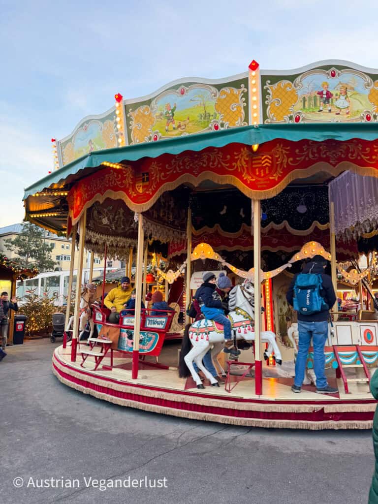 Vintage carousel with children riding painted horses and parents standing nearby, surrounded by festive decorations and holiday lights in a cheerful market setting.