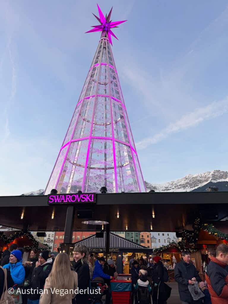 A towering Swarovski crystal Christmas tree glows in pinkish-purple hues at dusk, surrounded by a bustling crowd and wooden stalls with the Alps visible in the background.