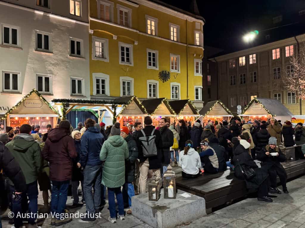 Crowds gather at night in front of a row of Christmas market stalls with white rooftops and string lights, set against yellow and white townhouses in Innsbruck’s Altstadt.