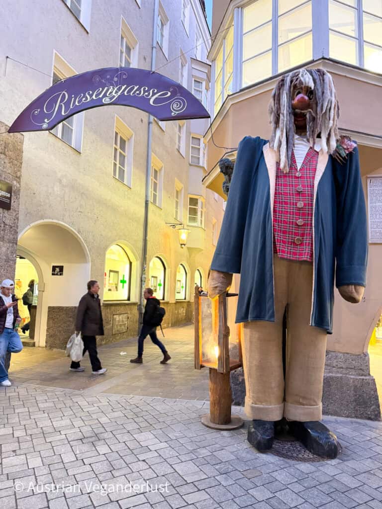 A towering figure of a giant with a red plaid vest, dreadlocks, and oversized clothes stands beside a lantern at the entrance to Riesengasse alley, part of Innsbruck’s fairy tale-themed holiday displays.