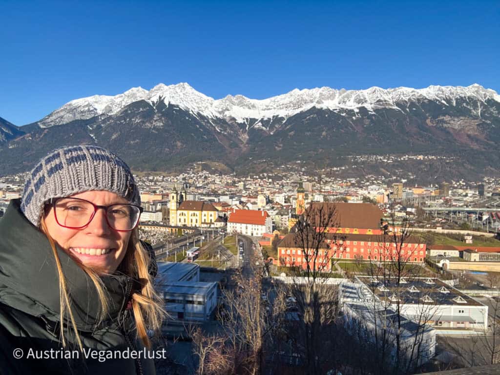 A smiling woman in winter clothing takes a selfie with Innsbruck’s cityscape and snowy mountains in the background, under a clear blue sky and bright sunlight.