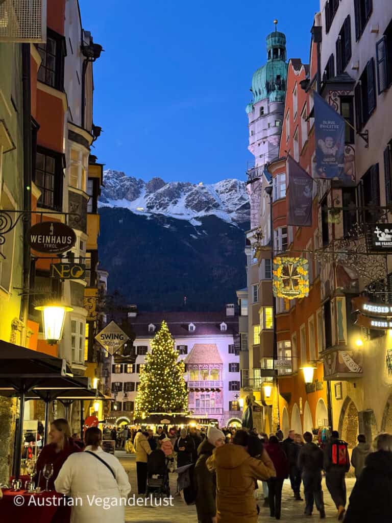 Evening view of Innsbruck's historic old town with a festive Christmas tree glowing in front of the Golden Roof, as people stroll between warmly lit buildings with the snow-capped Nordkette mountains rising in the background.