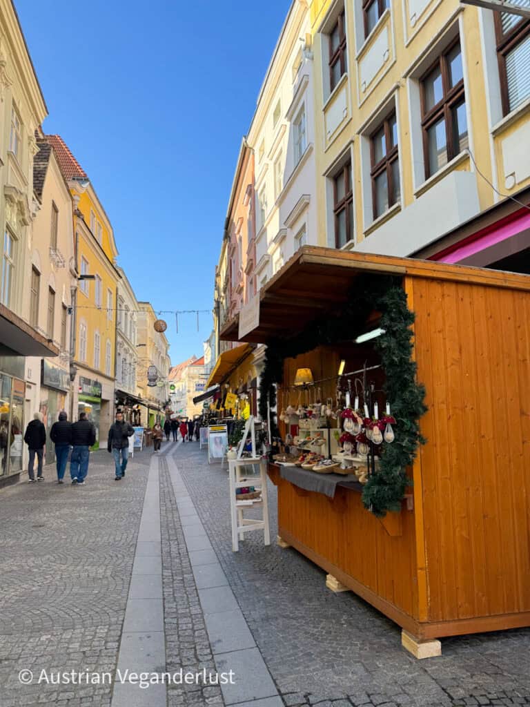 A cheerful pedestrian shopping street lined with pastel buildings and festive market stalls selling handmade ornaments and gifts. Locals stroll through the sunny, blue-skied Christmas market atmosphere.