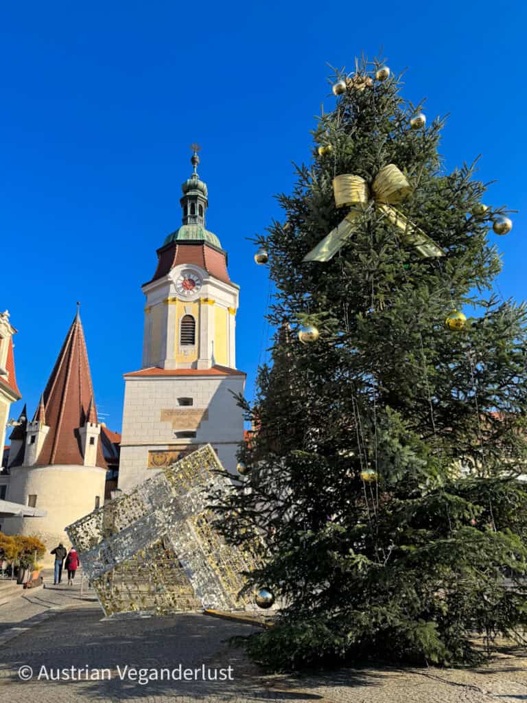 A tall Christmas tree decorated with gold ornaments and a large golden bow stands in a sunny town square near a tilted cube-shaped light sculpture, with a clock tower and historic buildings in the background under a clear blue sky.