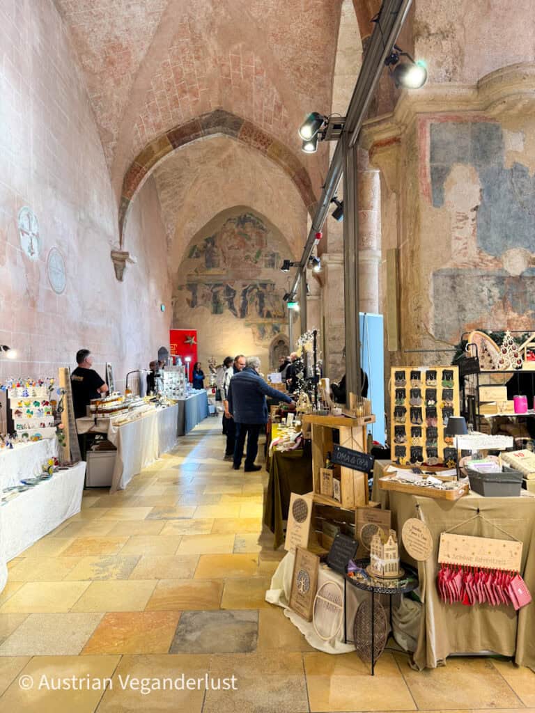 A cozy indoor Christmas market set inside a vaulted, medieval-style hall with faded frescoes on the stone walls. Tables are filled with handmade crafts, decorations, and gifts, with a few people browsing the stalls.