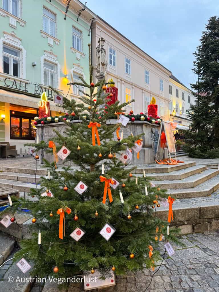 A Christmas tree decorated with orange ribbons, small baubles, and wish tags stands in front of a historic fountain with oversized red candle decorations, set against pastel buildings in a charming European square.