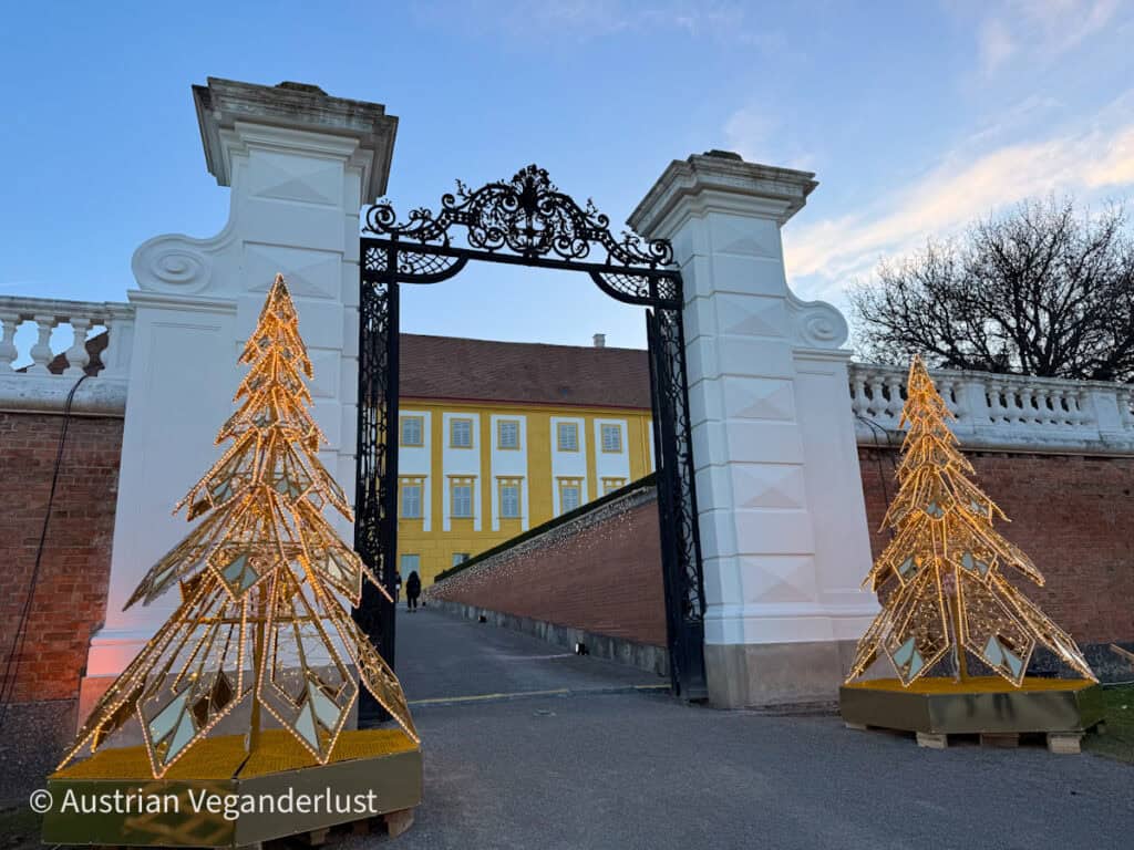 Two golden, light-adorned Christmas trees frame an ornate wrought iron gate leading into the courtyard of a yellow baroque building. The photo captures the entrance beautifully lit at dusk.