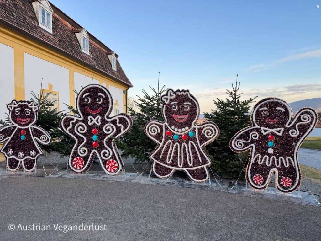 Four large gingerbread character decorations with smiling faces and colorful candy details stand in front of evergreen trees and a yellow building. The playful figures create a cheerful holiday scene in daylight.