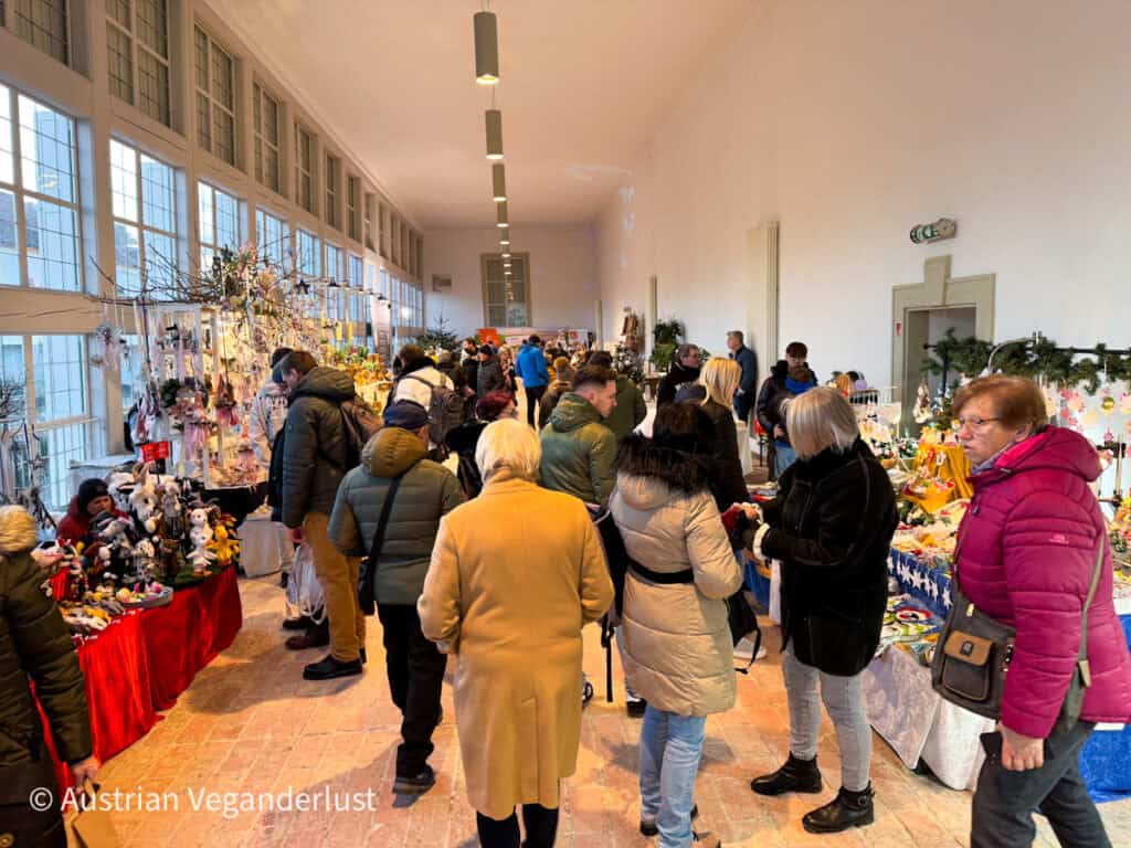 A bright indoor hall filled with people browsing handmade holiday crafts and decorations at various vendor tables. Shoppers in winter coats examine plush toys, ornaments, and festive goods under soft ceiling lights.