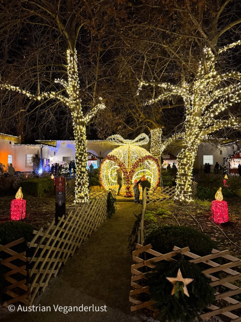 A festive garden path lined with wooden fencing and red candle decorations leads to a giant ornament-shaped light installation, glowing with golden and red lights under trees wrapped in fairy lights. Visitors are seen walking through the ornament tunnel at night.
