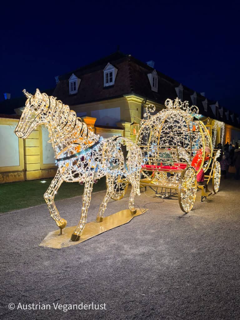 A stunning illuminated sculpture of a horse pulling an ornate carriage, both outlined in golden lights with red accents, stands against a baroque-style building at night. The scene evokes a fairytale atmosphere perfect for a holiday display.