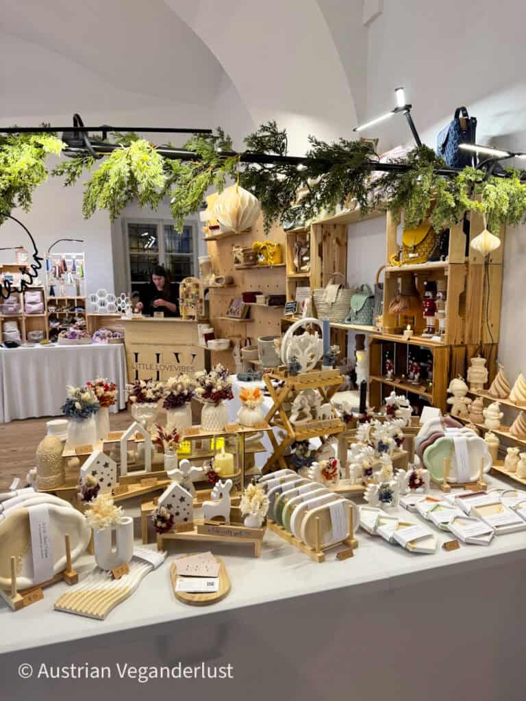 A beautifully arranged indoor Christmas market stall presents handmade ceramic and wooden items including vases with dried flowers, nativity figures, and decorative signs. The display is adorned with evergreen garland and soft lighting for a cozy feel.
