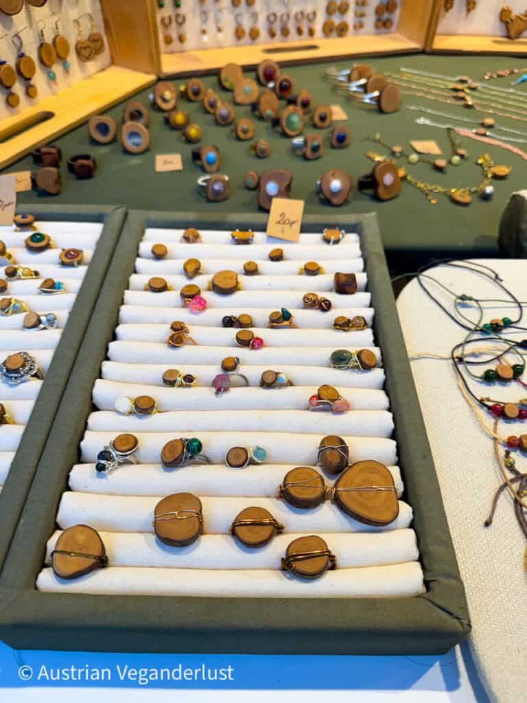 A jewelry vendor's table showcases handcrafted wooden rings, earrings, and pendants set with colorful stones and wrapped in wire. The rings are neatly organized in fabric trays, with price tags visible on some pieces.