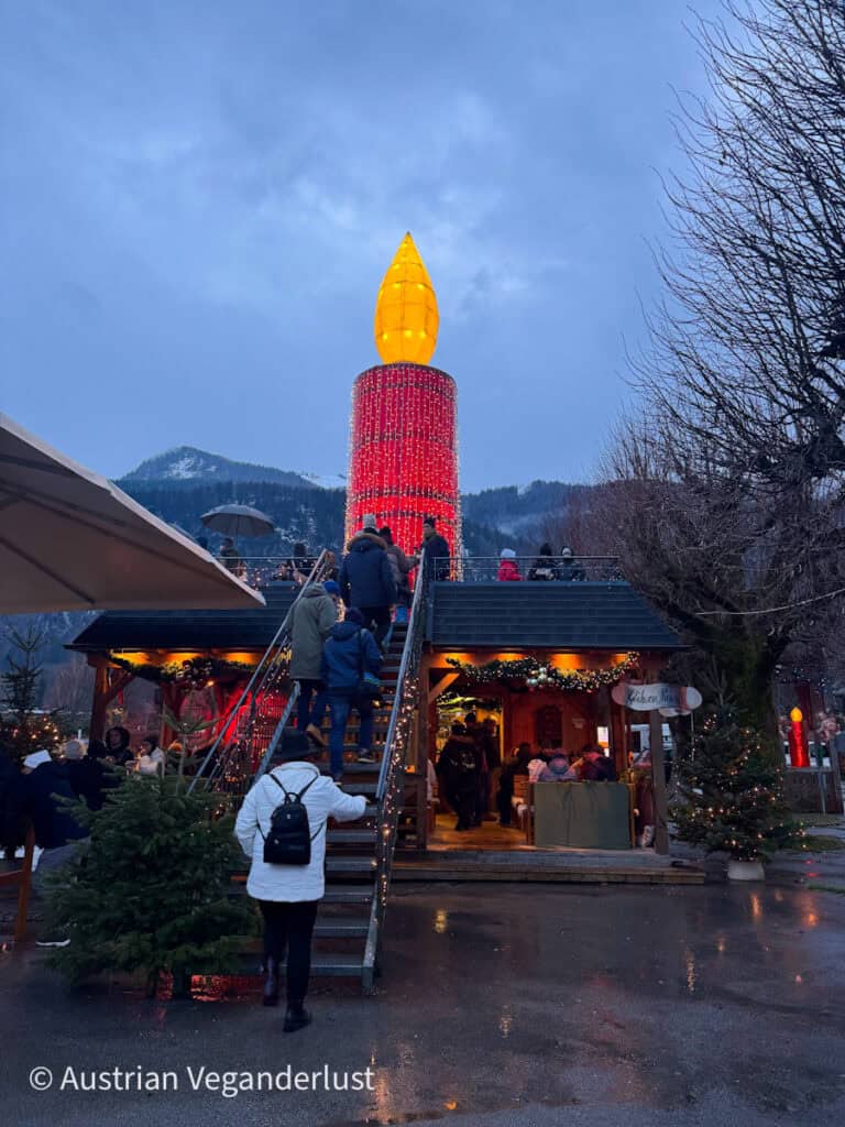 Crowds climb stairs leading to a platform beside a massive red candle lit with golden lights, surrounded by wooden huts and alpine scenery.
