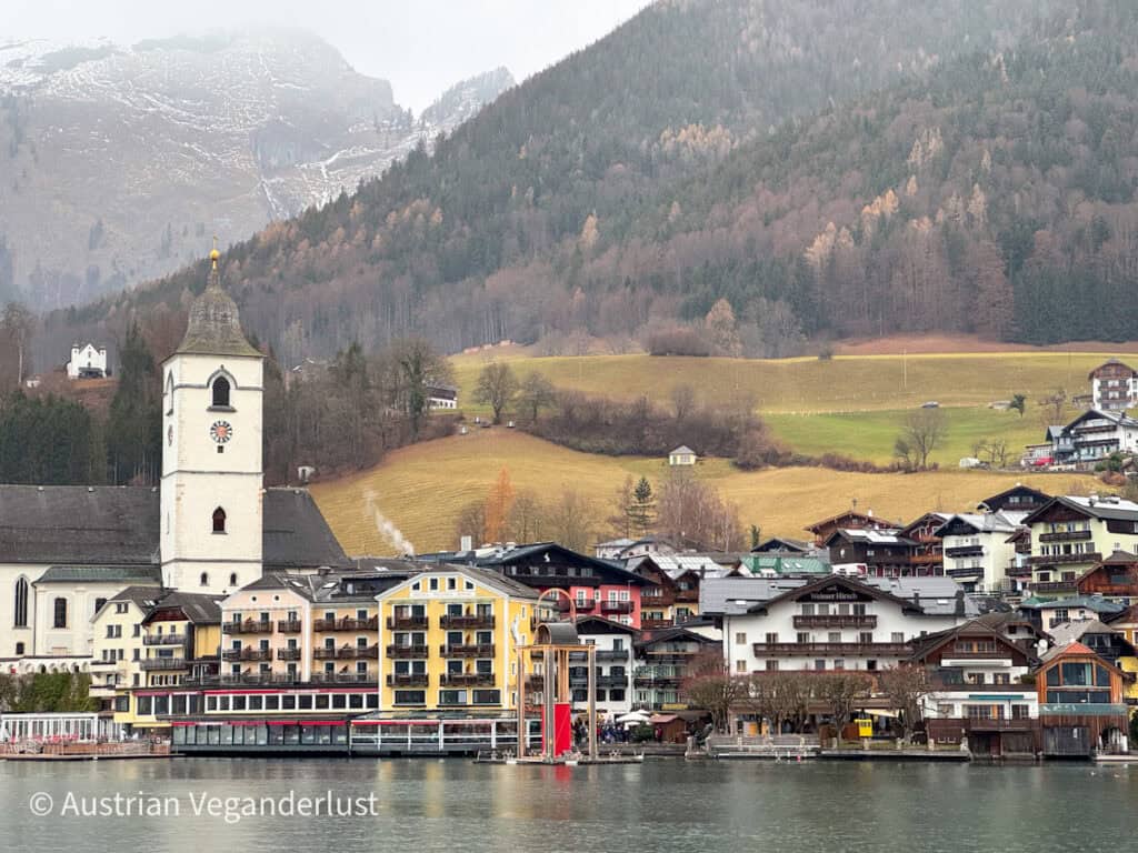 Daytime view of St. Wolfgang with its white church tower and colorful alpine buildings lining the lakefront, with snowy hills in the background.