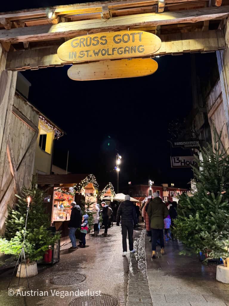 Visitors walk under a wooden sign reading “Grüß Gott in St. Wolfgang” at the entrance to the Christmas market, with lit stalls lining the narrow street.
