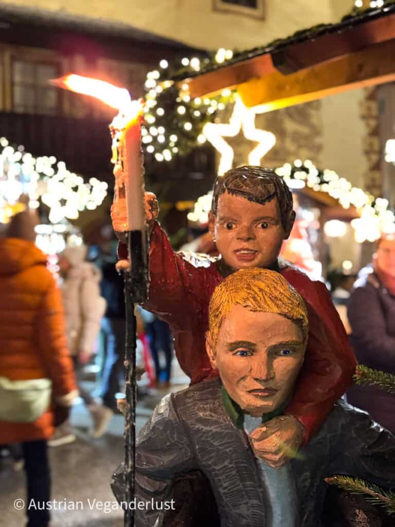 Close-up of a festive wooden statue showing a boy holding a candle while sitting on a man’s shoulders, surrounded by glowing star lights.