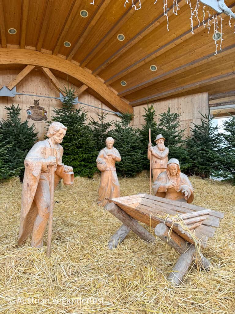 Full-sized hand-carved nativity scene in a straw-covered shelter surrounded by evergreen trees at the Strobl Christmas market.