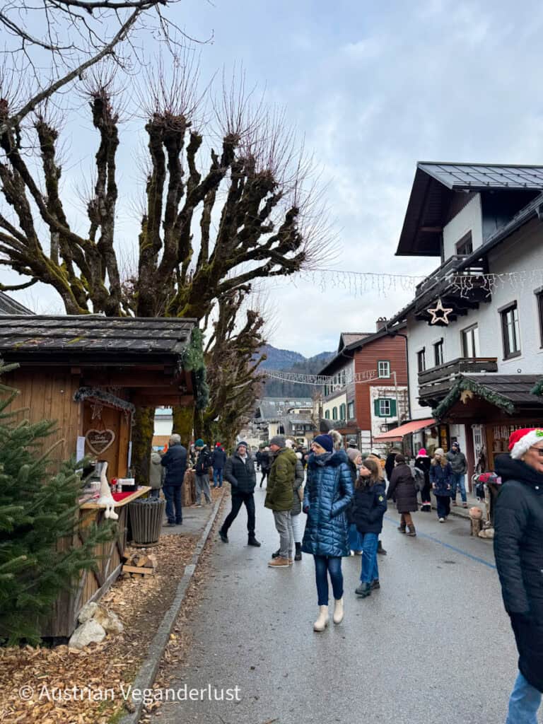 Visitors stroll along a festive street lined with bare, sculptural trees and wooden Christmas stalls in Strobl, with alpine buildings in the background.