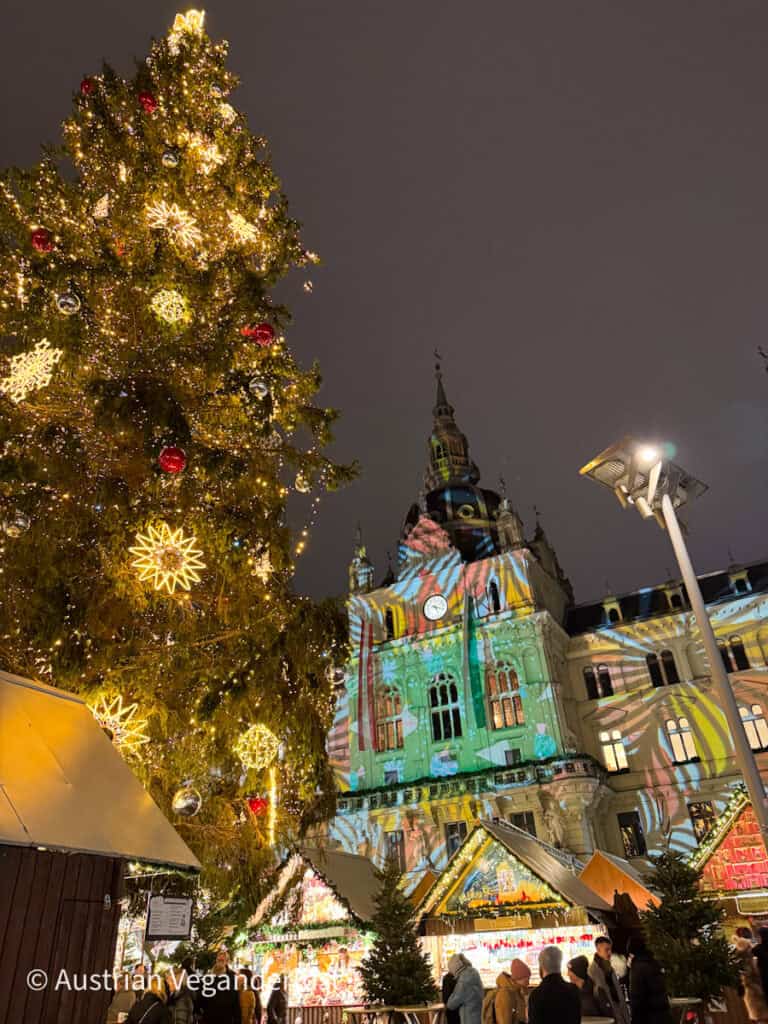 A massive Christmas tree adorned with lights and ornaments stands tall in front of Graz City Hall, which is illuminated with colorful holiday projections during a festive night market.