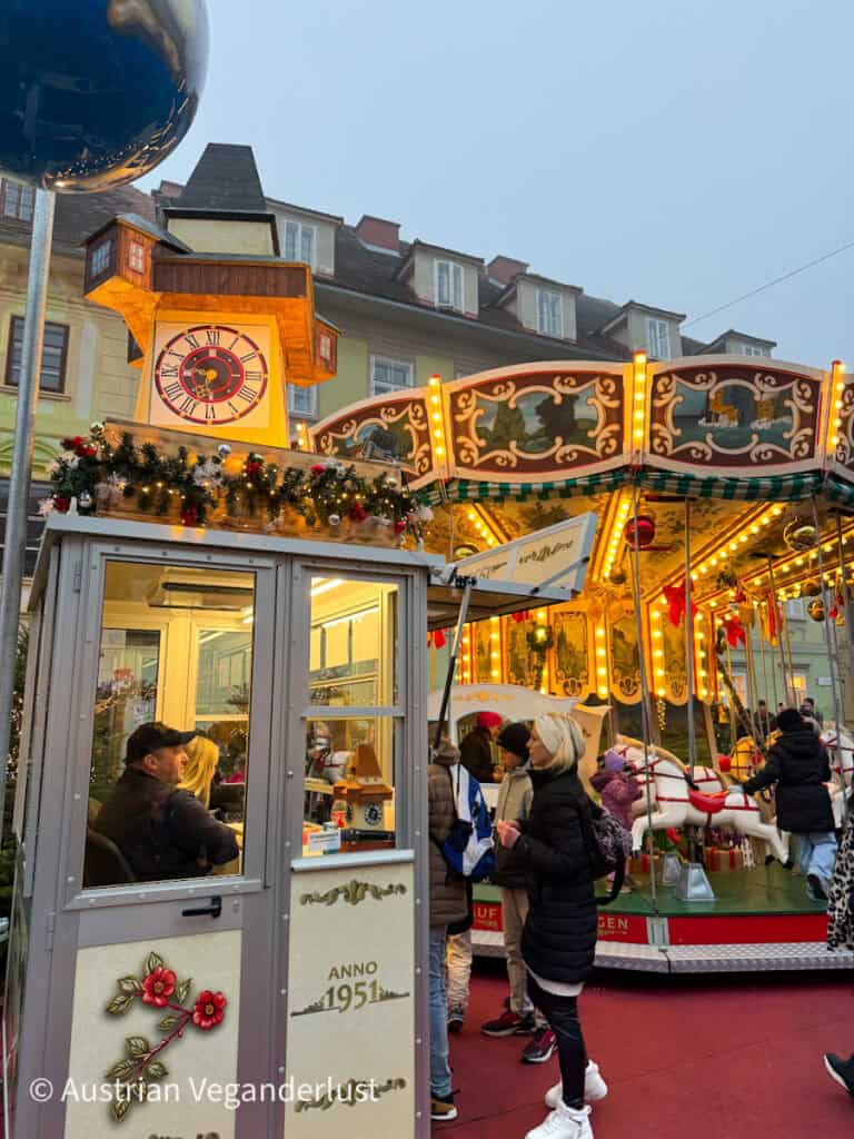 A cheerful Christmas market scene with a vintage-style carousel and a decorated ticket booth. People wait in line beside the carousel, which features white horses and ornate patterns, with a clock and garlands above.