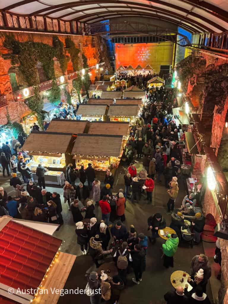 A bustling Christmas market inside a covered, rustic stone structure. Numerous decorated wooden stalls line the space, with crowds enjoying food, drinks, and shopping beneath festive lighting.