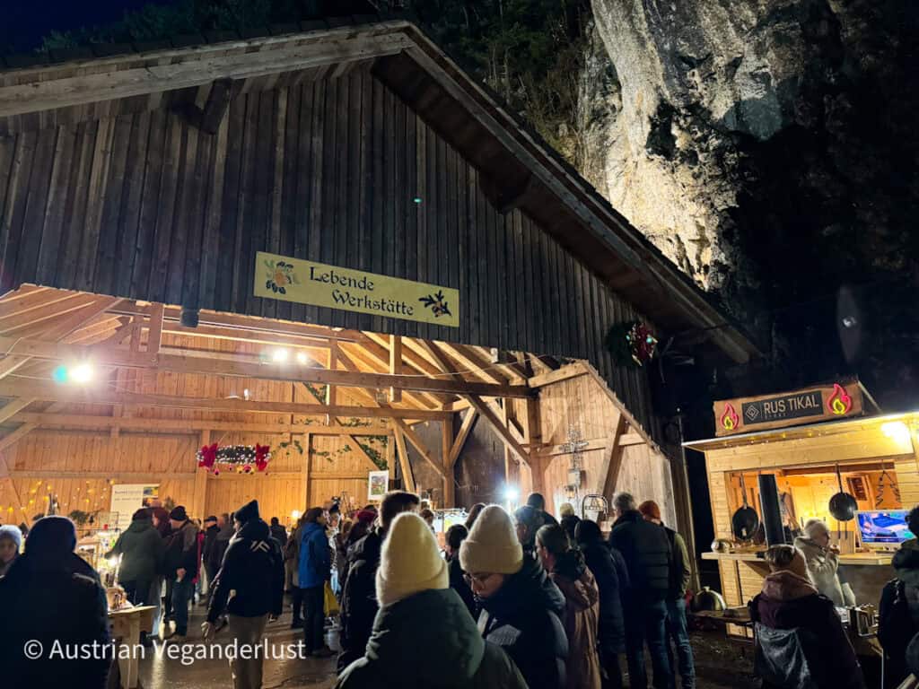Nighttime scene at a rustic wooden barn labeled "Lebende Werkstatt" (Living Workshop) bustling with visitors browsing crafts and artisan stalls; festive lights and cozy ambiance.