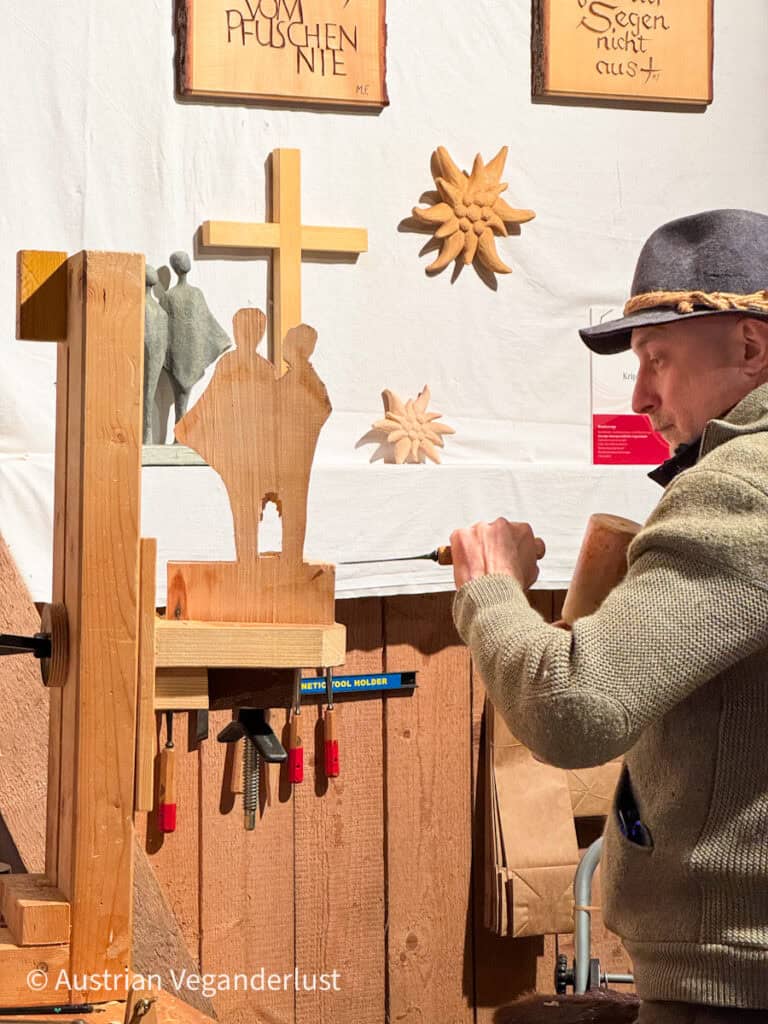 Close-up of a craftsman in traditional attire carving a nativity-themed wooden sculpture, surrounded by handcrafted wall decor and wood ornaments.