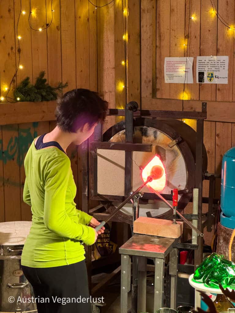 A woman in a neon green top shaping molten glass in a glowing furnace at a glassblowing workshop booth decorated with string lights.