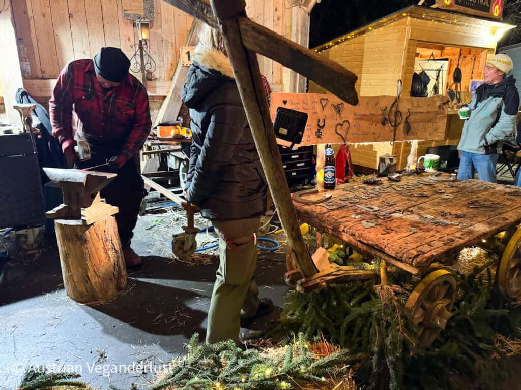 A blacksmith in a red plaid shirt works at an anvil while a woman watches; handmade iron crafts are displayed on a rustic wooden cart nearby.