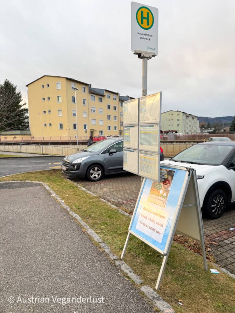 Bus stop sign at "Neunkirchen Bahnhof" with schedules posted and a board listing departure times; cars and apartment buildings in the background.