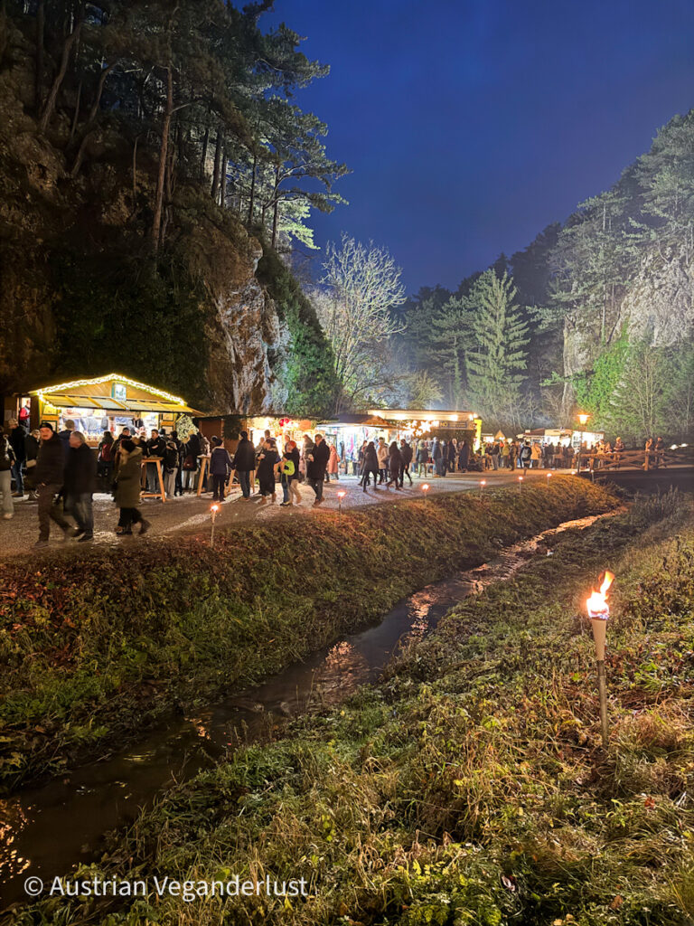 A lively Christmas market nestled between rocky cliffs and pine trees, with festive stalls lit up and crowds browsing under a deep blue evening sky.