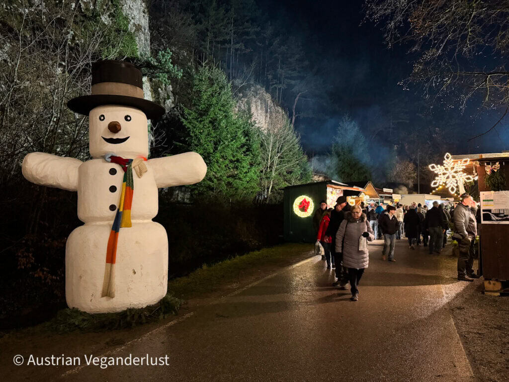 A large, smiling snowman sculpture with a colorful scarf greets visitors at the entrance of a Christmas market lined with glowing snowflake decorations and festive stalls.