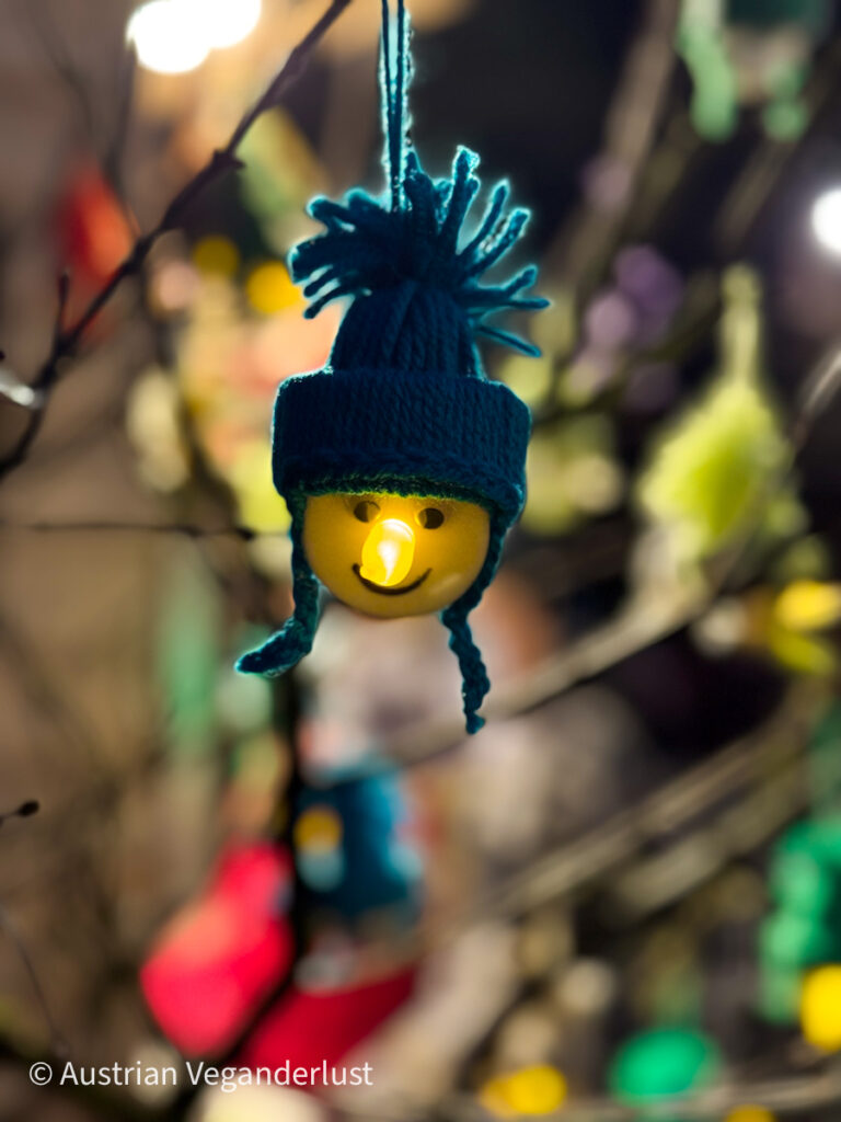 A close-up of a glowing smiley face ornament with a tiny light inside its nose, wearing a teal woolen winter hat, hanging from a tree branch in soft focus festive surroundings.