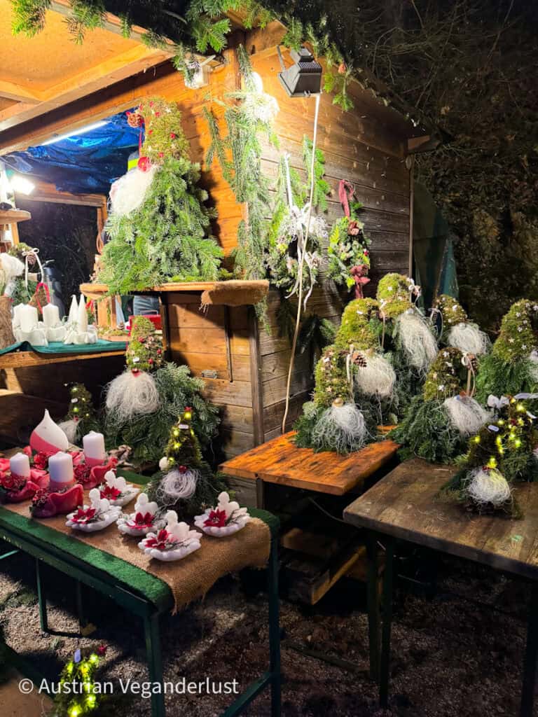 A rustic market stall adorned with evergreen decorations and filled with handmade Christmas crafts like gnome trees, candles, and festive centerpieces made from natural materials.