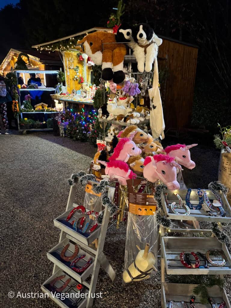 A playful vendor booth at the Christmas market displaying stuffed animals, plush unicorns, and painted horseshoes for sale, all under festive lights with more stalls in the background.