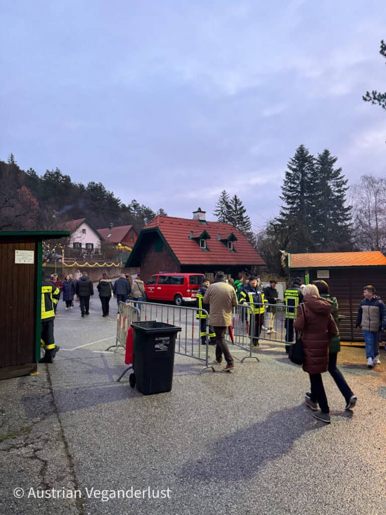 Entry point to the market with people walking through a checkpoint manned by firefighters; cozy houses with red roofs in the background.