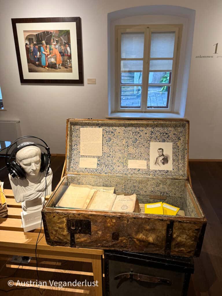 Ein alter, offener Koffer, ausgekleidet mit Blumenpapier, in dem alte Dokumente, ein kleines Porträt und Informationskarten zu sehen sind, neben einer weißen Büste mit Kopfhörern in einem Museumsraum mit klassischer Einrichtung.