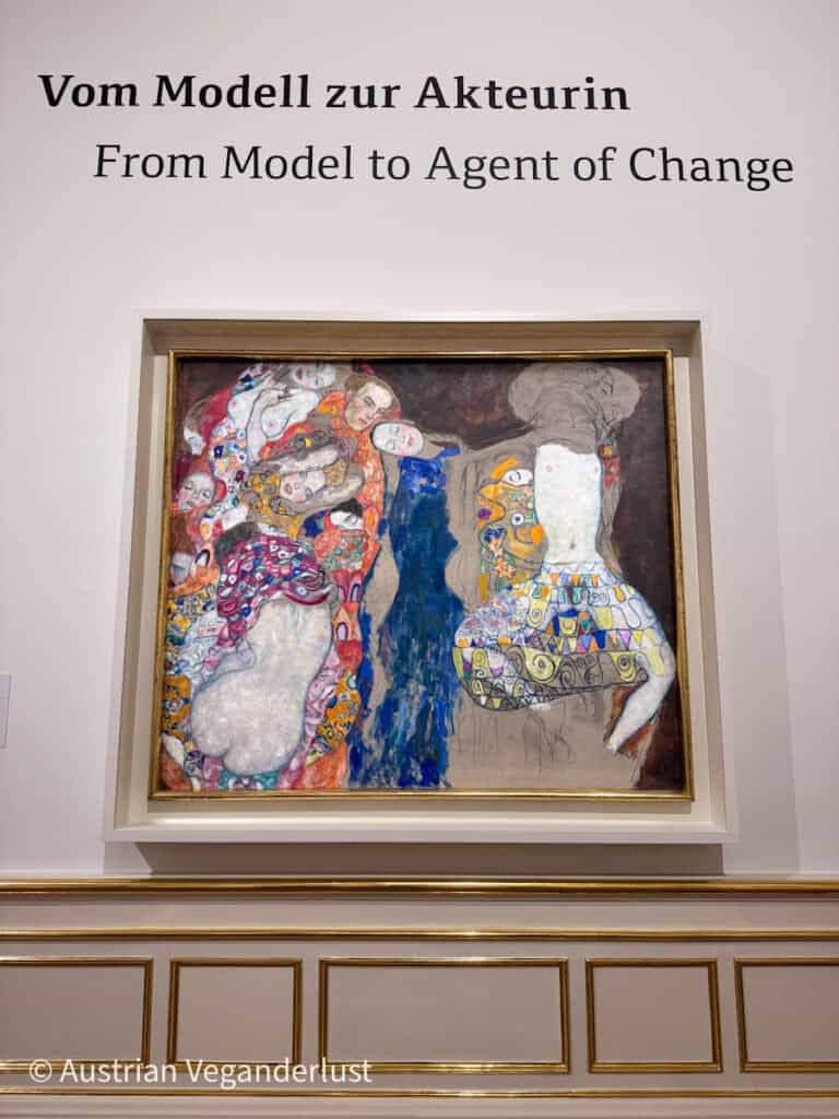 A large, vibrant painting depicting intertwined figures in richly patterned garments, with bold colors including red, blue, and gold. Above the artwork, wall text reads “Vom Modell zur Akteurin – From Model to Agent of Change.” The painting is displayed in a gold frame within a white gallery mount.