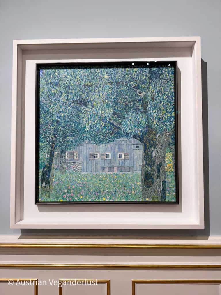 A landscape painting showing a small wooden house nestled among trees in a lush, flower-filled meadow. The scene is rendered in dense, textured brushstrokes of green, blue, and yellow. The artwork is framed in black and set within a white mount on a museum wall.