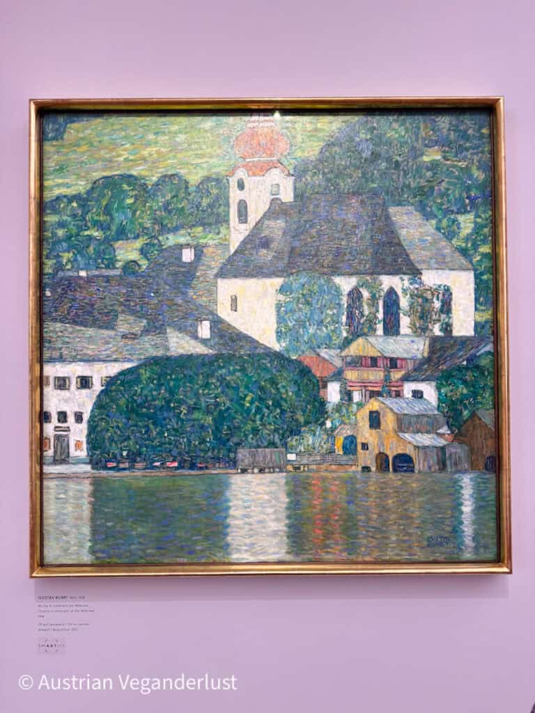 Ein gerahmtes Landschaftsgemälde von Gustav Klimt, das einen Kirchenkomplex am Seeufer zeigt, dessen weißer Turm von einer roten Kuppel gekrönt wird. Eng beieinander stehende Häuser und üppig grüne Bäume säumen das Ufer, deren Farben sich in der plätschernden Wasseroberfläche spiegeln. Die Szene ist in sanften, mosaikartigen Pinselstrichen mit überlagerten Grün-, Blau- und Goldtönen dargestellt.