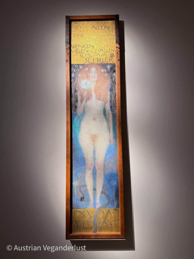Gustav Klimt’s Nuda Veritas shows a full length nude woman with long red hair standing against a deep blue background and holding a round mirror in her raised hand. A dark serpent coils at her feet and a gold panel above her displays a German quote reading Kannst du nicht allen gefallen durch deine That und dein Kunstwerk mach es wenigen recht. Vielen gefallen ist schlimm. Schiller. The tall narrow painting is framed in dark wood with the words Nuda Veritas at the bottom.