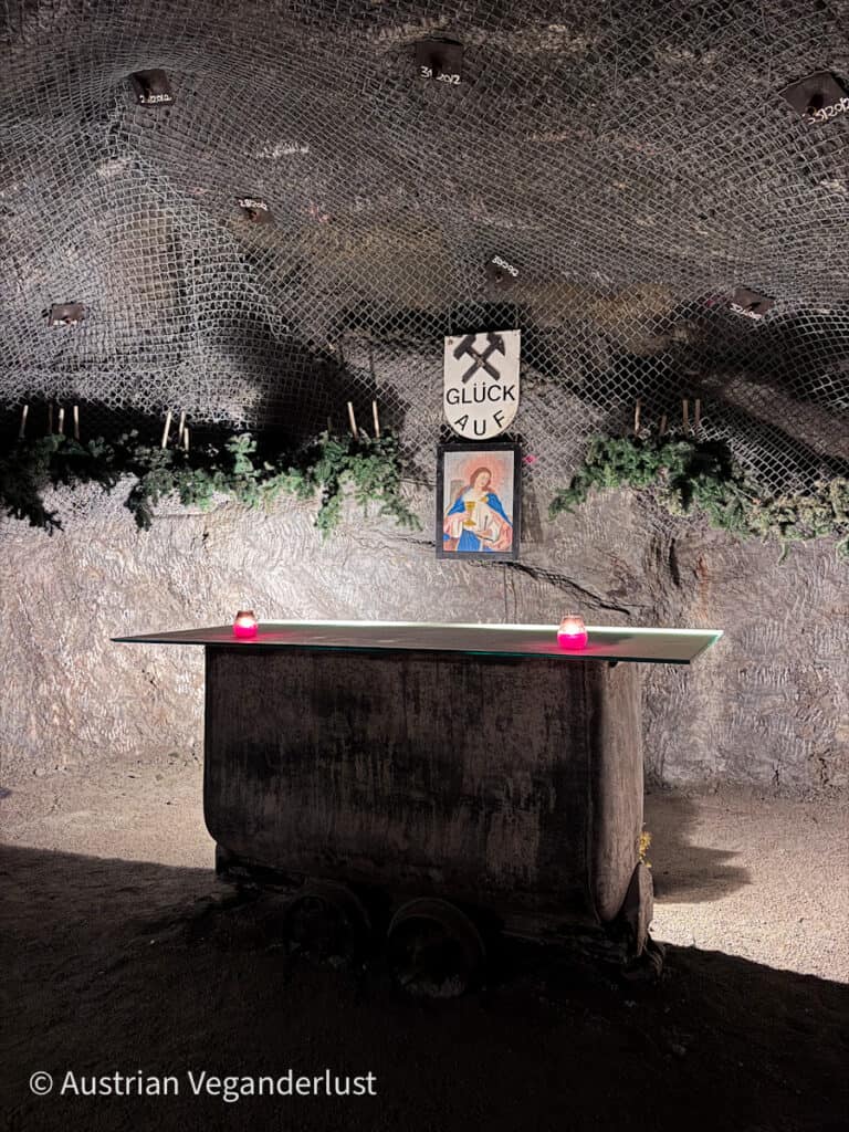 Ein schlichter unterirdischer Altar auf einem Grubenwagen, geschmückt mit Kerzen, immergrünen Zweigen und einem religiösen Bild, unter einem Schild mit der Aufschrift „Glück Auf“ in einem befestigten Stollen.