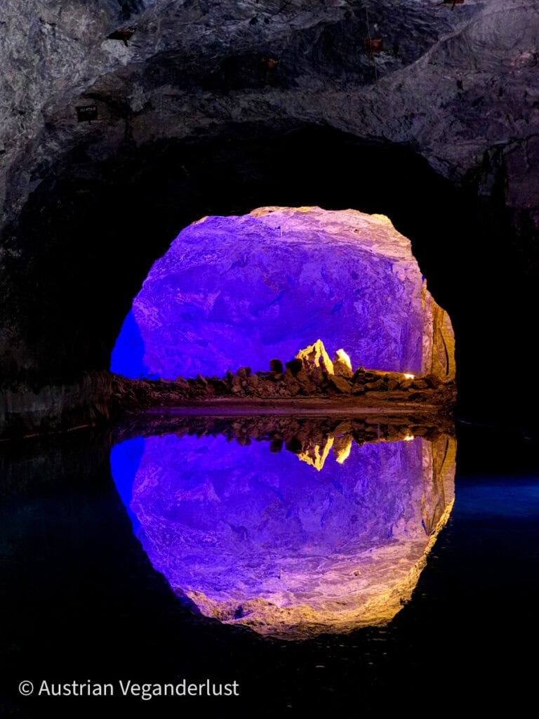 Eine große, beleuchtete Felsformation, die sich perfekt im unterirdischen See der Seegrotte Hinterbrühl spiegelt und in der dunklen Höhle in leuchtenden Violett- und Goldtönen erstrahlt.